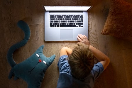 View from above a young child laying on floor watching videos on a laptop with a stuffed toy cat on floor next to child which has a skeptical side-eye facial expression, looking towards the child and computer