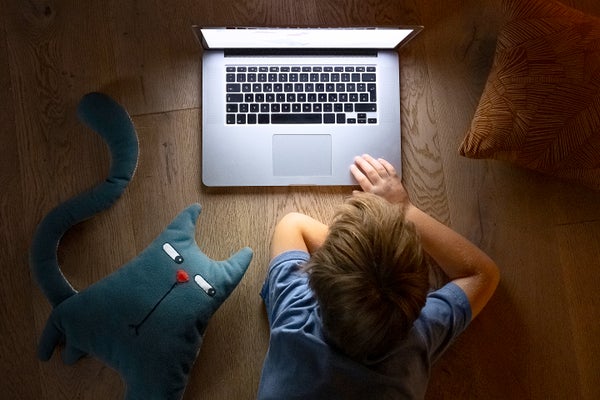 View from above a young child laying on floor watching videos on a laptop with a stuffed toy cat on floor next to child which has a skeptical side-eye facial expression, looking towards the child and computer