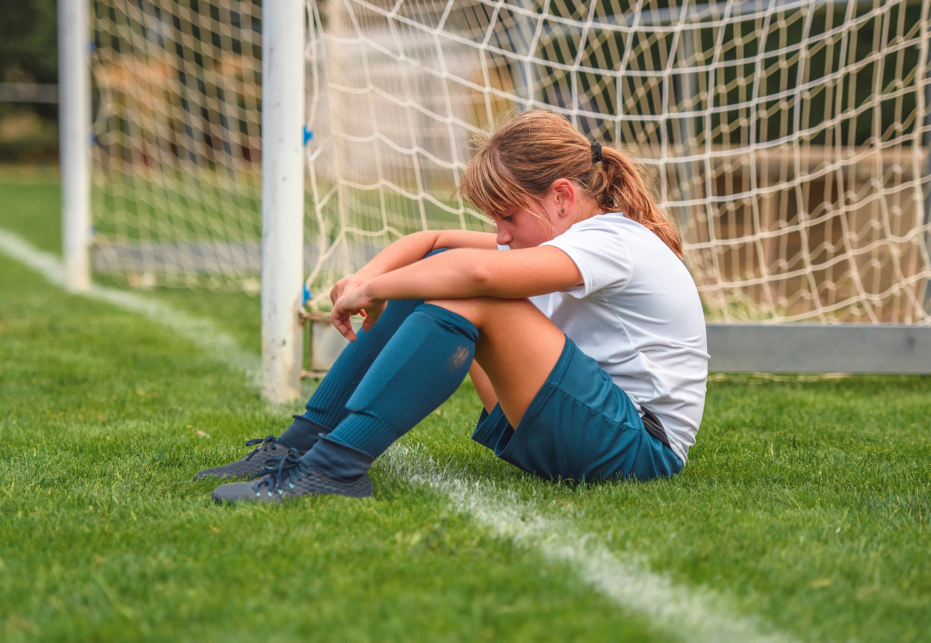 Side view of tired 9 year old female soccer player sitting on field next to goal thinking about how she played in practice.