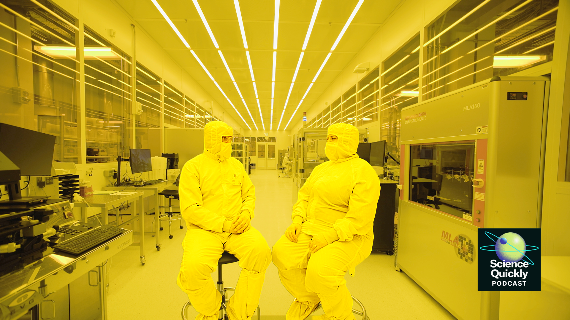 Two people in full clean room bunny suits sit on stools facing each other in a large yellow-colored room